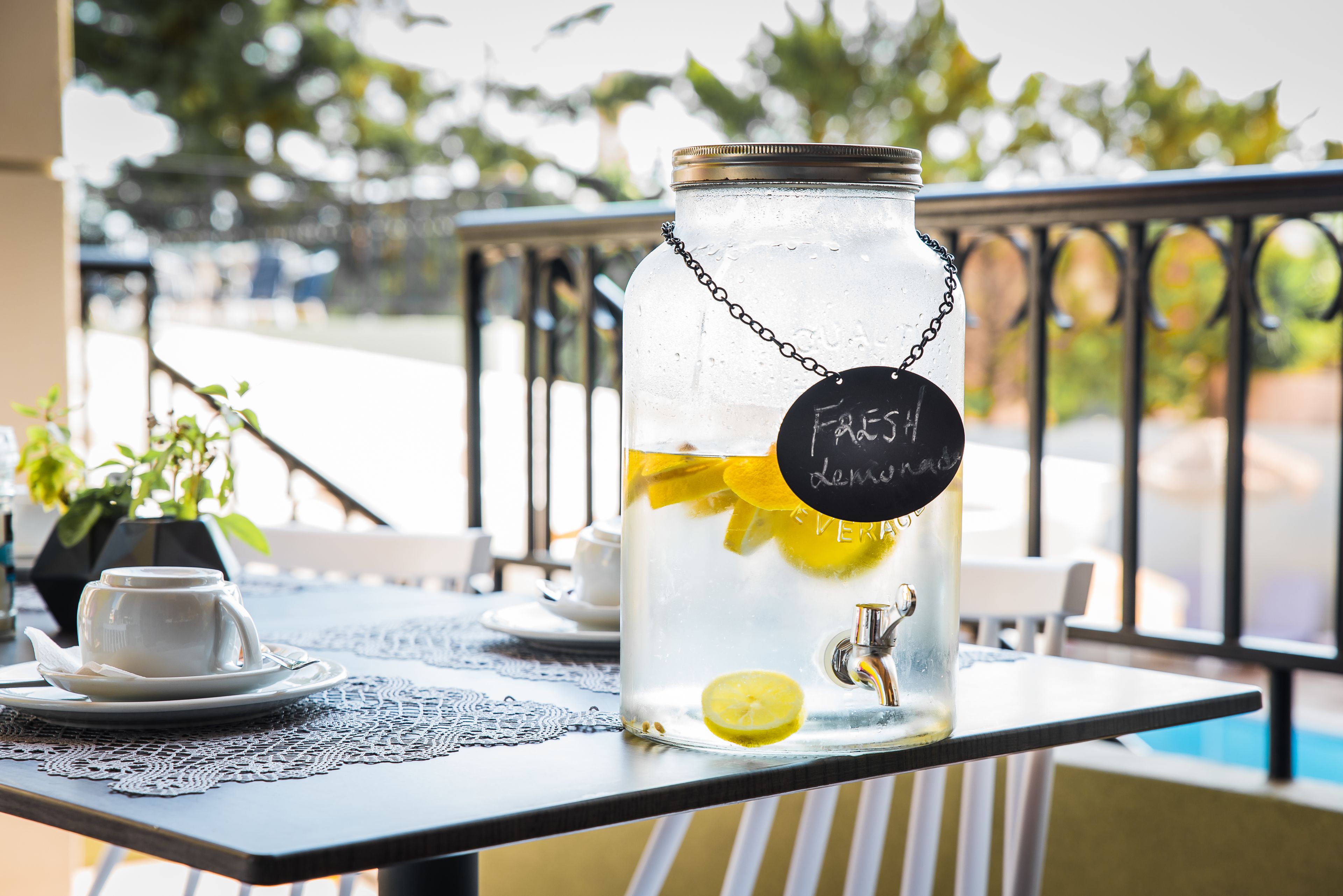 A glass jar with a metal lid and a chain, labeled "Fresh Lemonade," containing lemon slices and a lemon wedge, placed on a table with a lace doily.