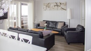 A modern living room with a black leather sofa set, a wooden coffee table, and a white dining table with chairs. There is a large window with curtains, a wall-mounted TV, and a floor lamp. - Hafjell Resort Hafjelltoppen Gaiastova (Øyer)