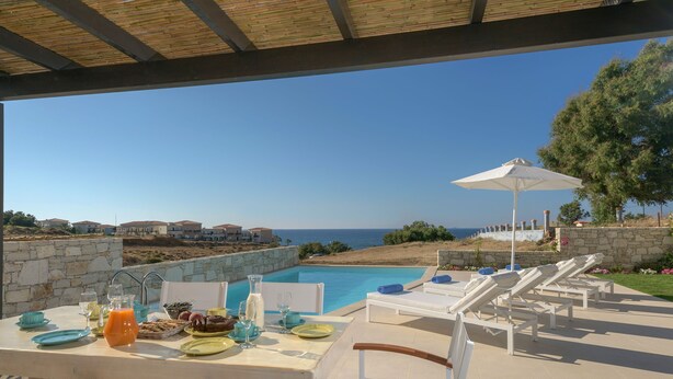A poolside dining area with a view of the sea, a table set with plates and glasses, and lounge chairs with umbrellas. - New Villa Louisa by the sea, walking distance from sea and shops, no car needed! (Πάνορμος)