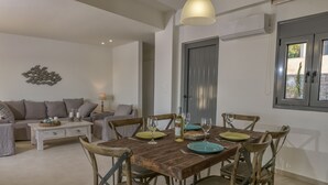 A dining area with a wooden table set for four, surrounded by rustic chairs. A white sofa and a small side table with a lamp are in the background. - New Villa Louisa by the sea, walking distance from sea and shops, no car needed! (Πάνορμος)