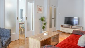 Un salon moderne avec une table basse en bois, un canapé recouvert d’un tissu à motifs rouges et noirs, un téléviseur posé sur un meuble blanc et une plante en pot.