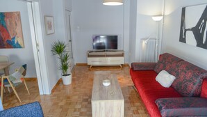 Un salon moderne avec une table basse en bois, un canapé rouge, un fauteuil bleu, un téléviseur posé sur un meuble en bois et une plante en pot.