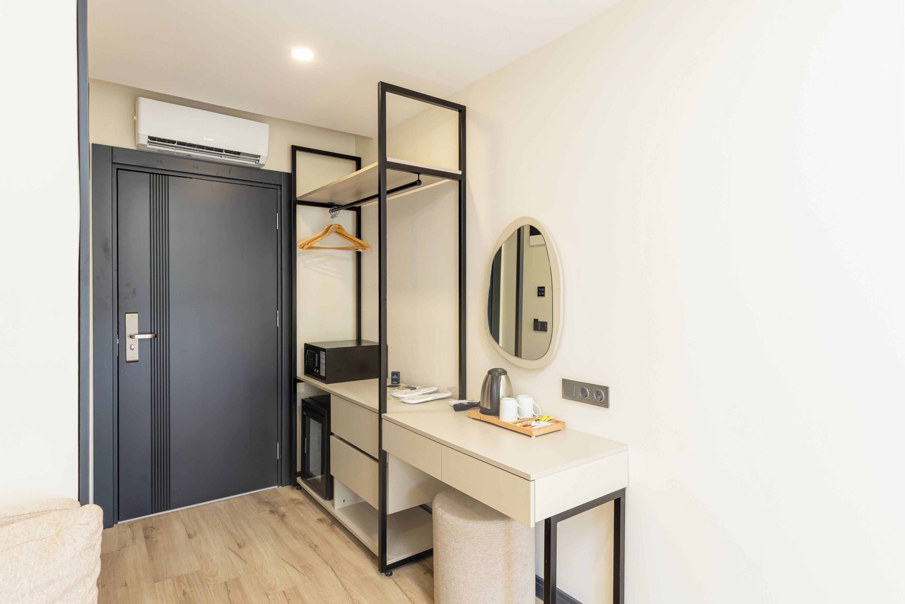 A compact hotel room with a desk, a wardrobe, a mirror, and a hanging coat rack.