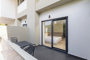 Un balcon avec une table et des chaises, une porte-fenêtre coulissante donnant sur une chambre à coucher équipée d’un lit et d’une fenêtre.