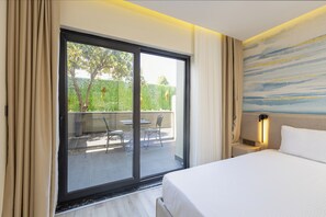 Une chambre d’hôtel avec un lit, une table de chevet, une lampe et une porte-fenêtre coulissante donnant sur un espace extérieur aménagé avec des chaises et une table.