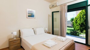 A neatly made bed with white linens and a folded towel, a bedside table with a lamp, and a window with a view of greenery. - Eleanna villas. Villa Athena (Καβρός)