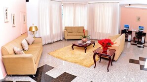Una sala de estar con un sofá color crema, una mesa de centro y una alfombra. Hay dos ventanas con cortinas blancas, un escritorio con una computadora y una silla.