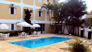 Una piscina con sillas y sombrillas blancas, rodeada por un edificio de paredes amarillas y múltiples ventanas.