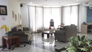 Un salón con sofá, sillones, una mesa de centro y televisor. Hay cortinas, una planta y un cuadro en la pared.