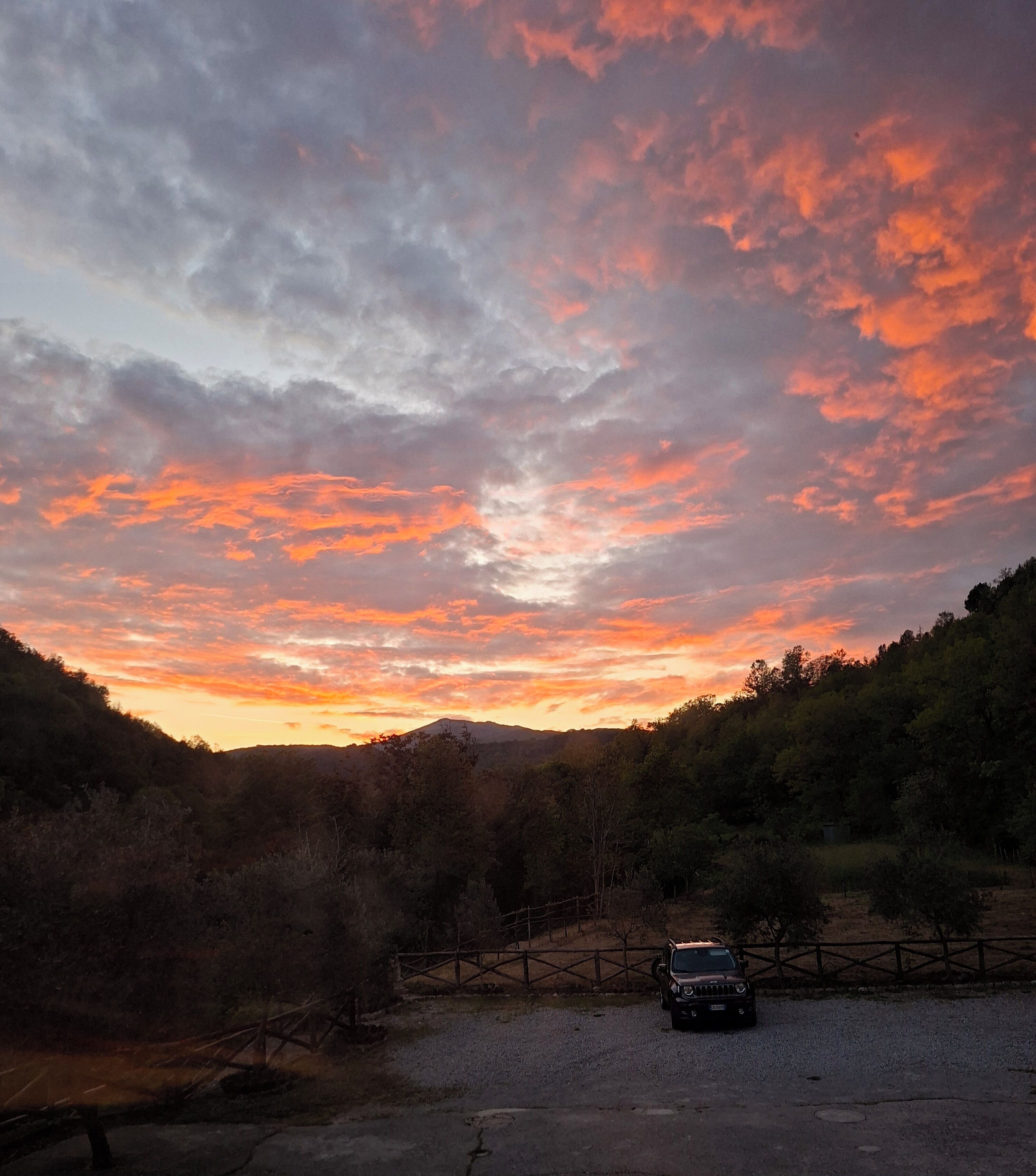 A gravel parking area with a vehicle, surrounded by a fence and trees, under a sunset sky.
