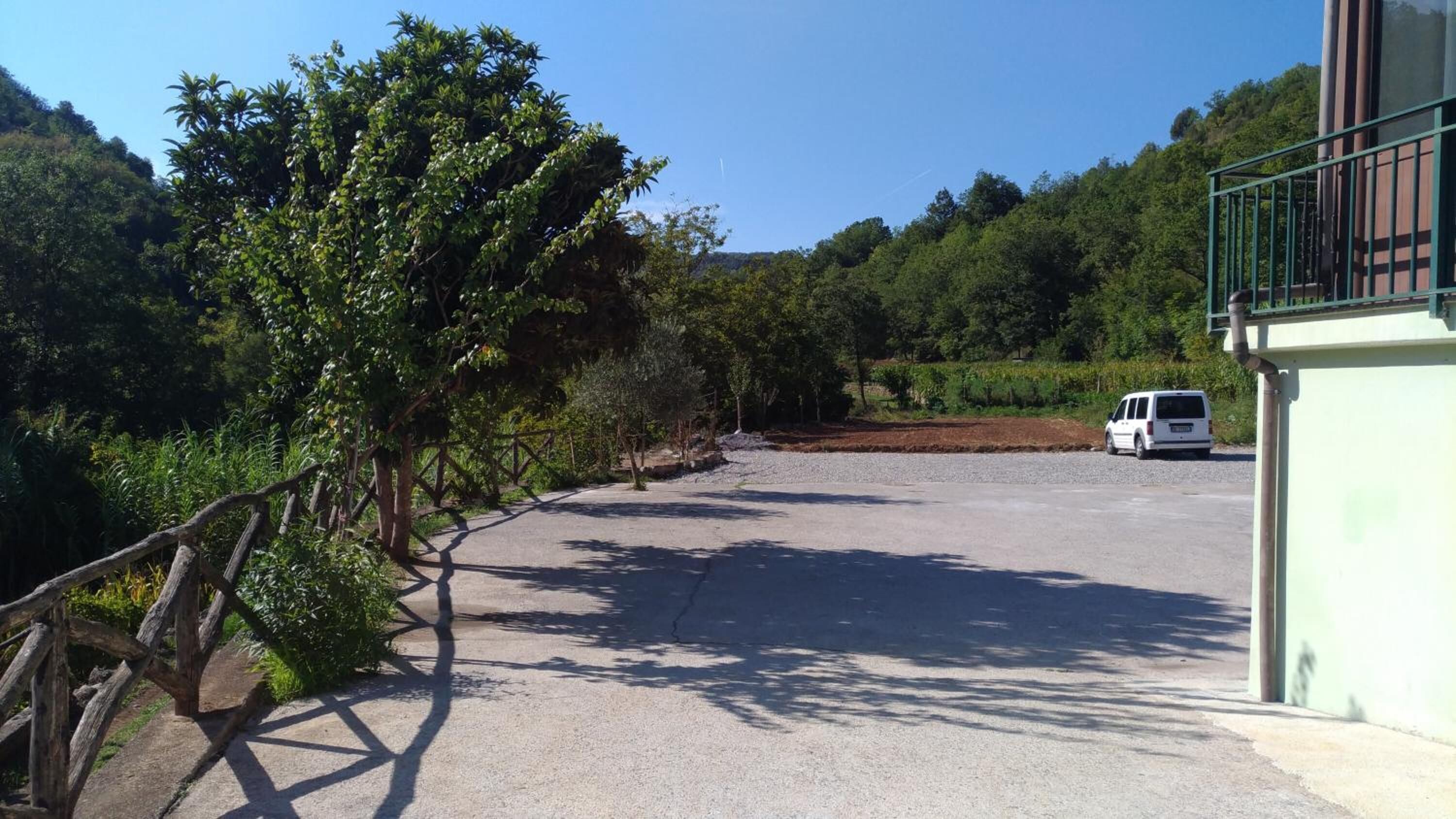 Uma área de estacionamento com uma carrinha branca, rodeada por árvores e um edifício com grades verdes.