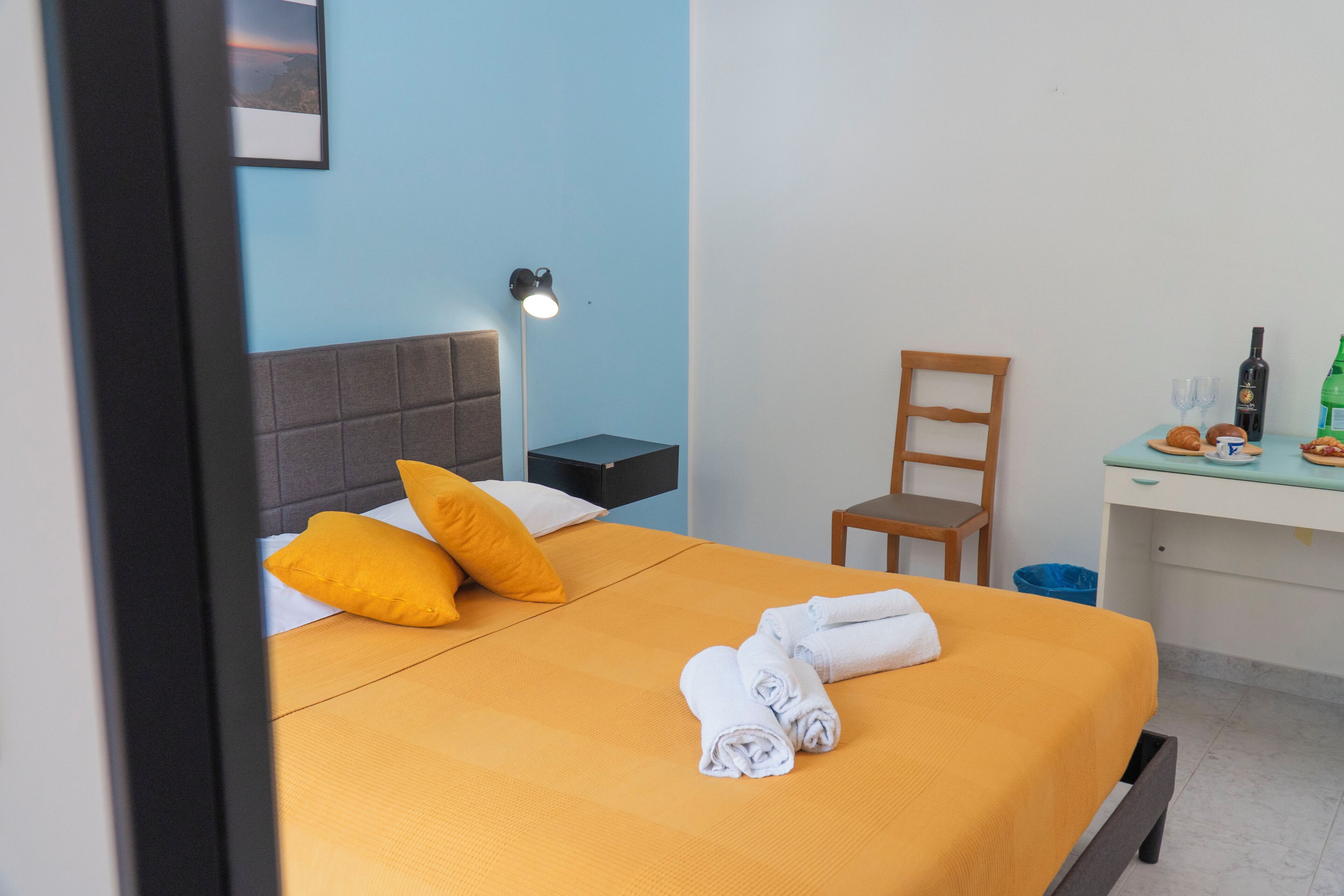 Una cama bien hecha con una colcha amarilla, dos almohadas y toallas enrolladas. Hay una mesita de noche con una lámpara, una silla y una pequeña mesa con una botella y vasos.