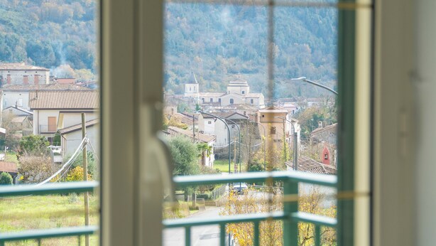 A view through a window of a town with buildings, a church, and a hill in the background. - Palia's Hotel (Laino Borgo)
