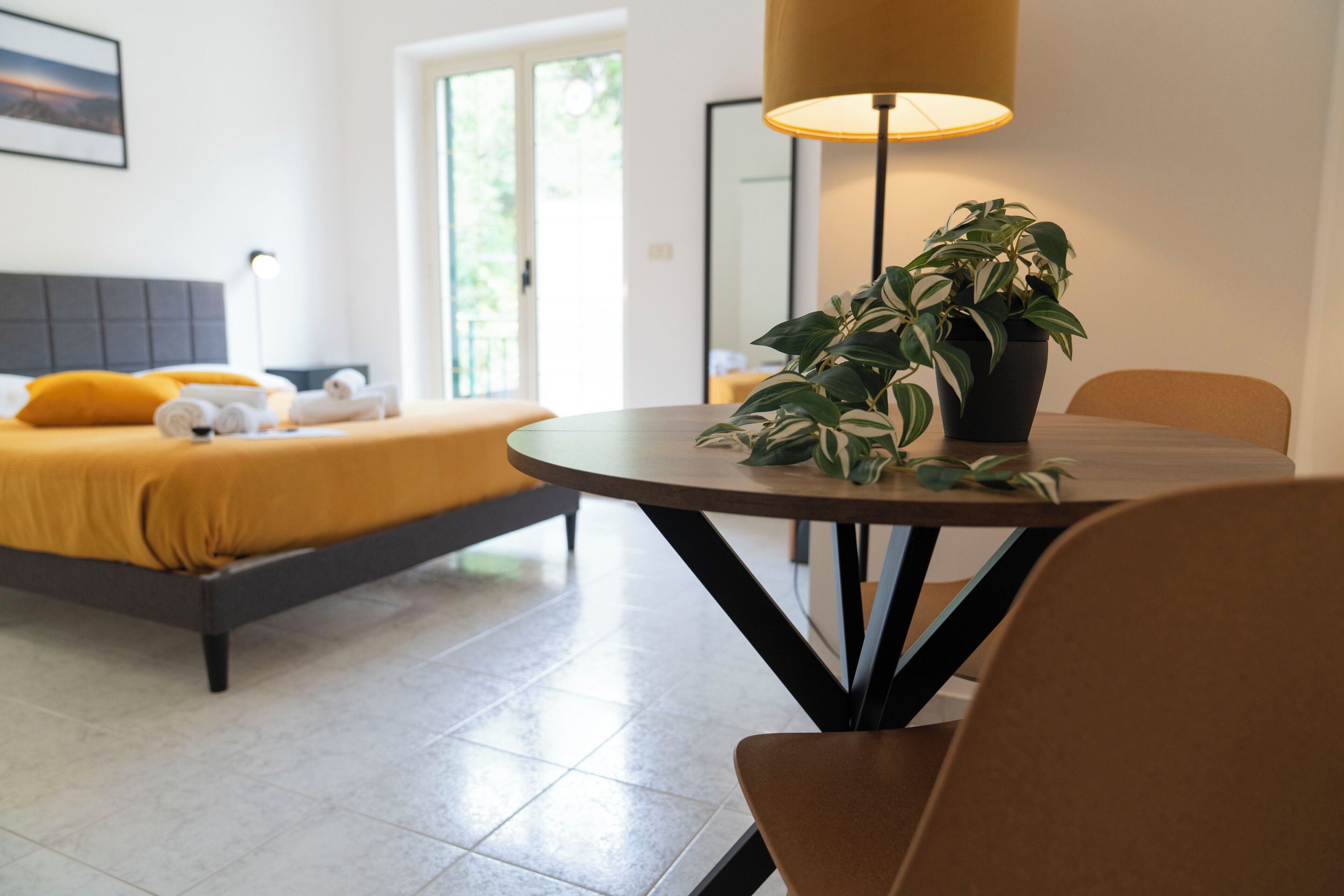 Una habitación de hotel con una cama, una mesa redonda con una lámpara y una planta en maceta.