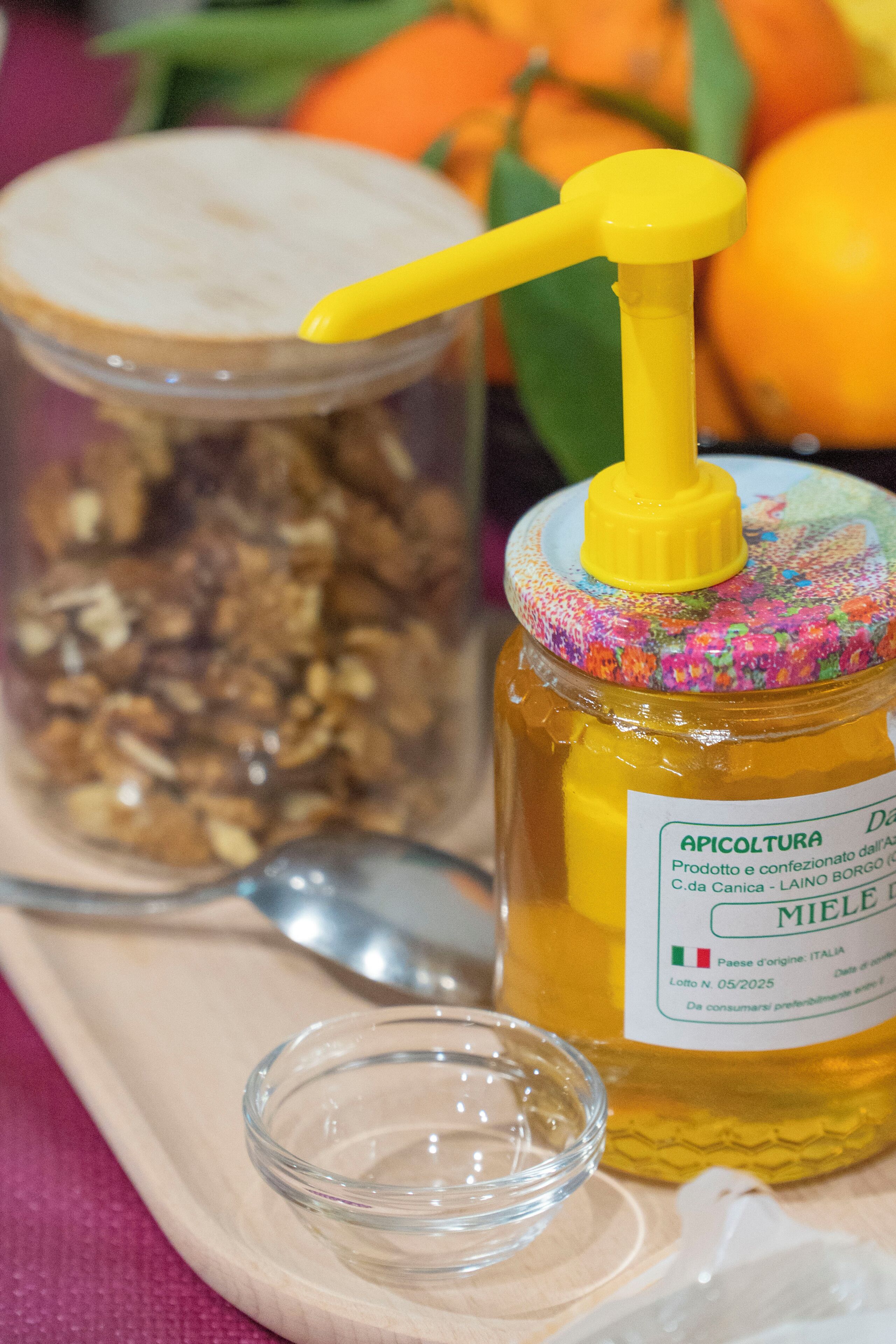 A jar of honey with a yellow dispenser, a glass of honey, and a container of granola on a wooden tray.