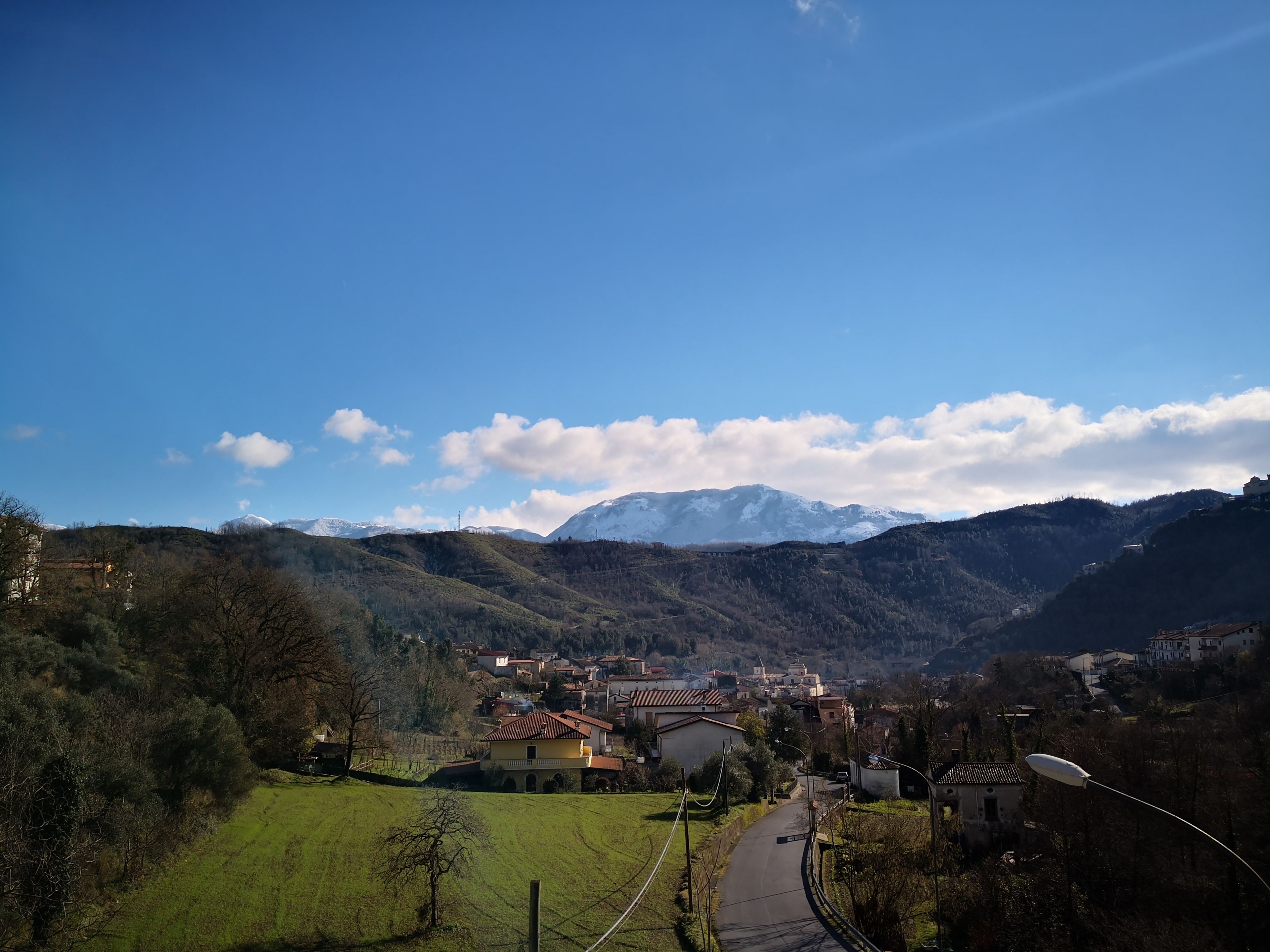 Un borgo rurale con case, una strada sinuosa e montagne innevate sullo sfondo.