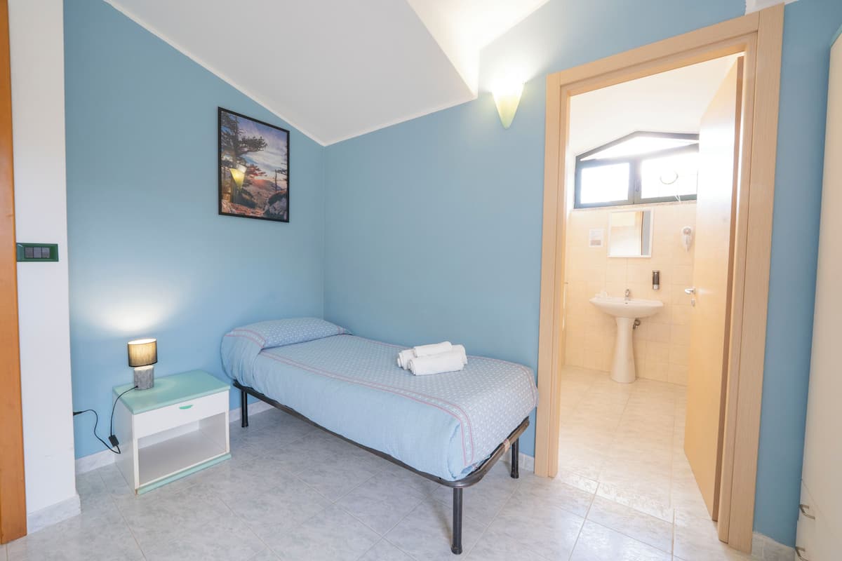Een slaapkamer met een enkel bed, nachtkastje, lamp en een schilderij aan de muur.