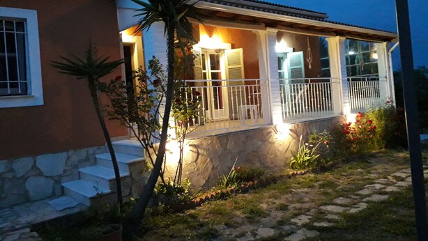 Ein Haus mit steinernen Fundament, einer Veranda mit weißen Geländern und einem kleinen Garten mit einer Palme und Topfpflanzen.
