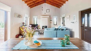 A spacious living room with a high wooden ceiling, a blue sofa, a dining table with a striped tablecloth, and a vase with flowers.