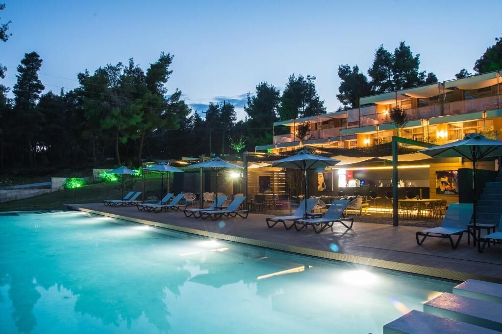 Une piscine avec des chaises longues et des parasols, un espace bar et un immeuble à plusieurs étages doté d’un éclairage extérieur.