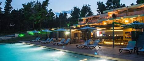 Une piscine avec des chaises longues et des parasols, un espace bar et un immeuble à plusieurs étages doté d’un éclairage extérieur.