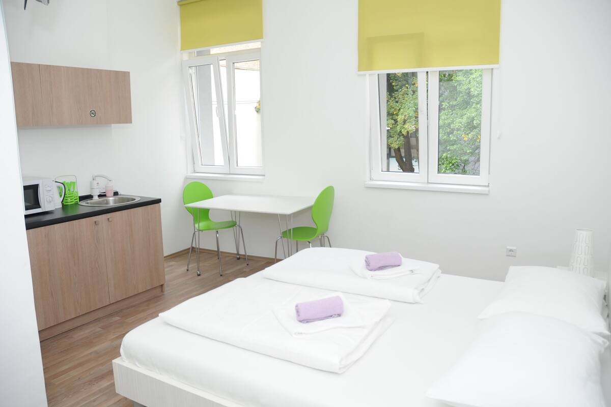 A modern hotel room with a bed, a desk, and a chair. There is a window with a view of trees.