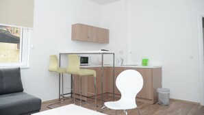 A modern, minimalistic room with a grey sofa, a white chair, a small kitchen area with a microwave, and a wooden cabinet.