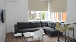 A modern living room with a sectional sofa, a small table, and two chairs. There are large windows with beige blinds, and a TV mounted on the wall.