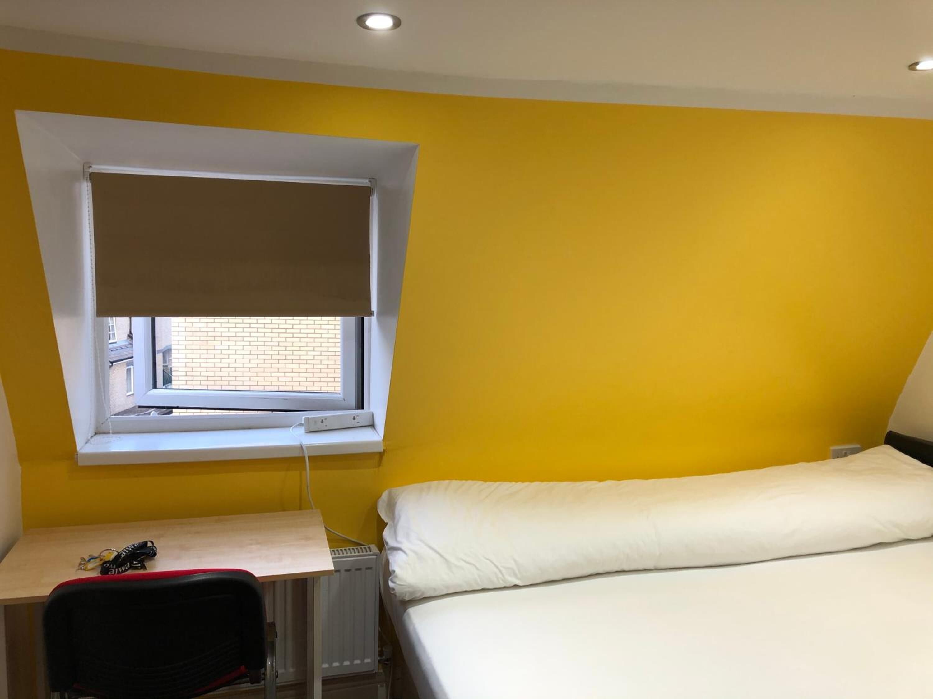 Une pièce aux murs jaunes, avec un lit, un bureau et une chaise, une fenêtre avec un store et un radiateur.