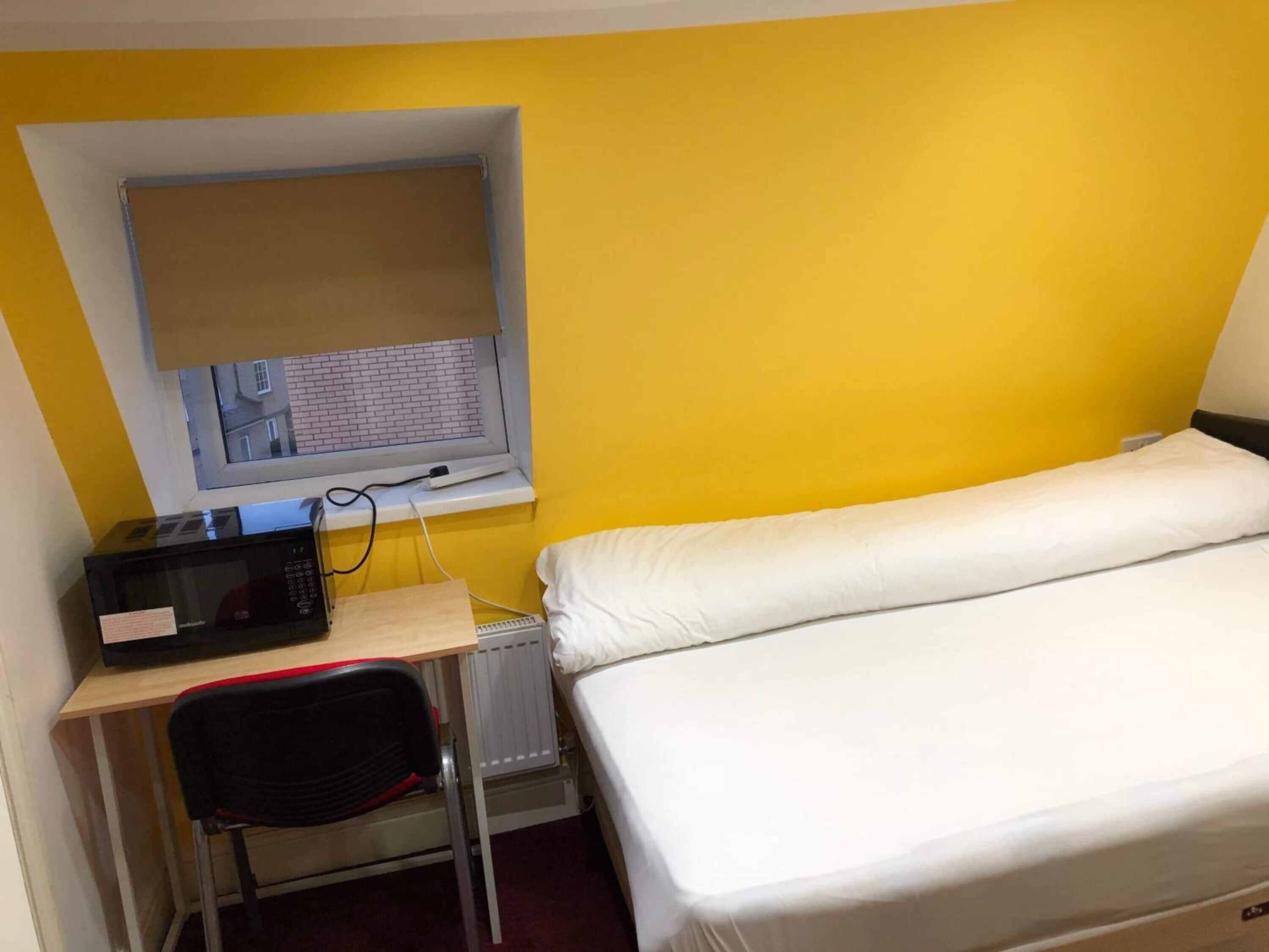 A small room with a bed, a desk, a chair, a window, and a microwave.