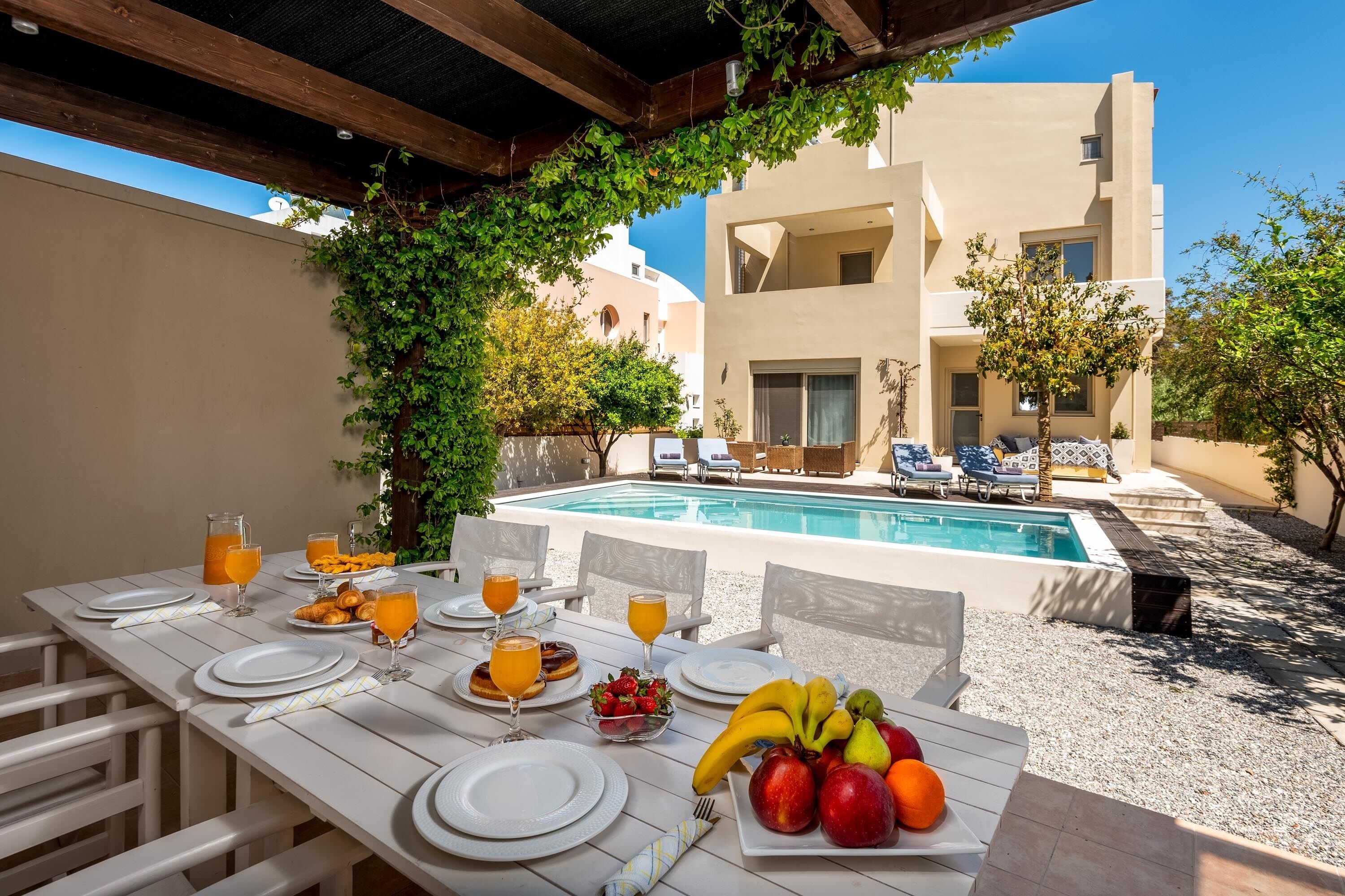 Mesa de jantar posta com pratos, copos e frutas, com vista para uma piscina e um edifício de design moderno.