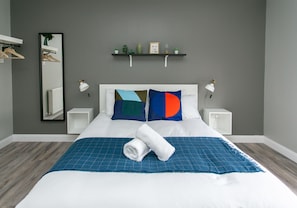 A neatly made bed with a blue checkered bedspread and a rolled white towel, two pillows with geometric patterns, a bedside table with a lamp, and a shelf with decorative items above the bed. - University Road Apartments (Belfast)