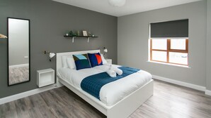 A modern bedroom with a white bed, grey walls, a large window, and a small shelf with decorative items. - University Road Apartments (Belfast)