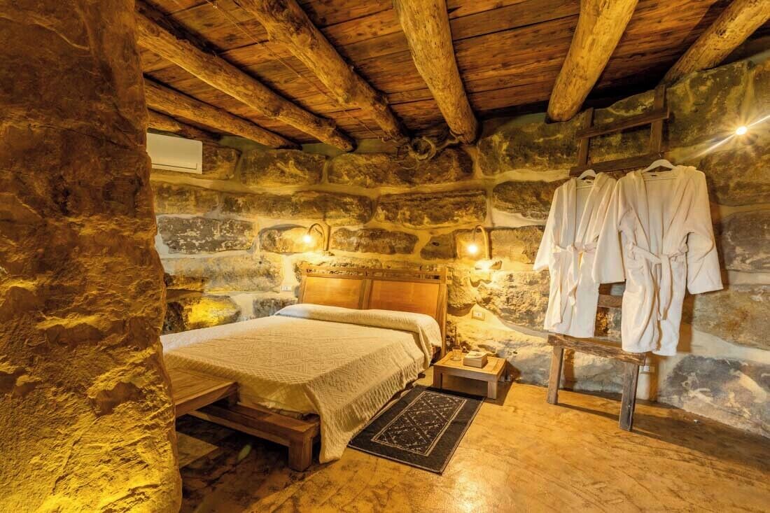 A rustic stone-walled bedroom with a wooden bed, a small bedside table, and a robe rack with robes.