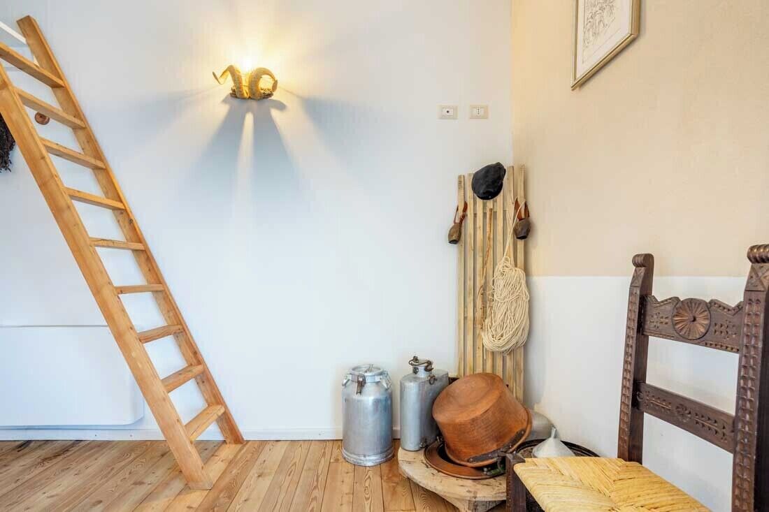 Une pièce avec une échelle en bois appuyée contre un mur blanc, un luminaire fixé au mur, deux récipients en métal, un chapeau en bois posé sur une chaise, et une chaise en bois dotée d’un dossier décoratif.