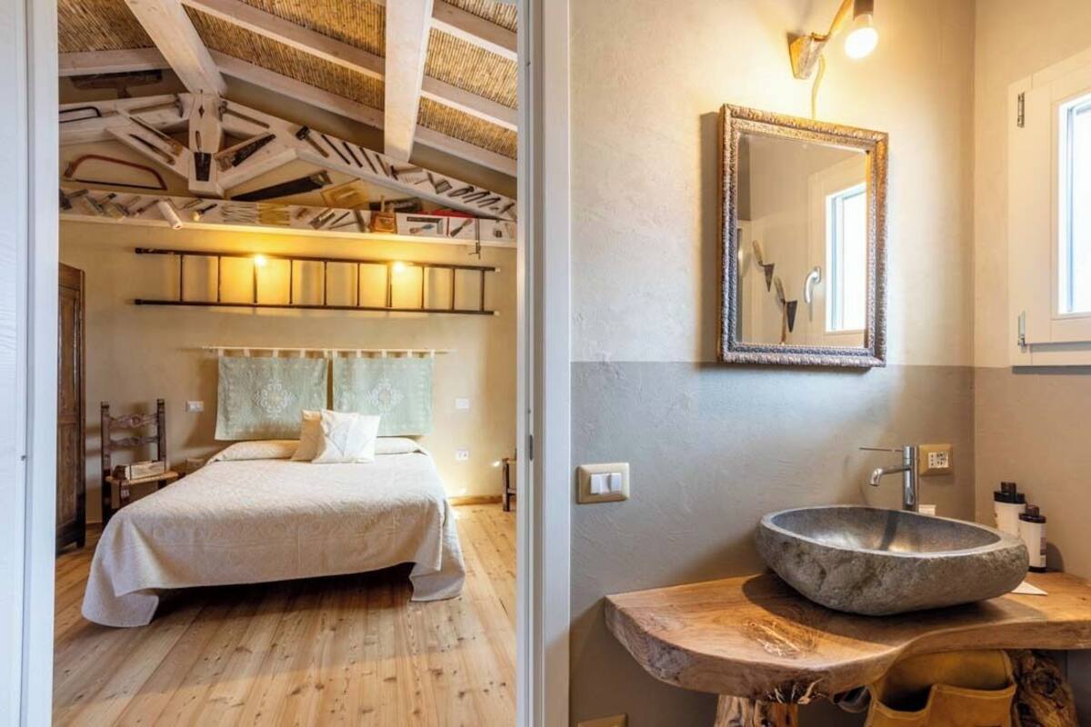 Ein Schlafzimmer mit Bett, Holzboden und Decke mit freiliegenden Balken. Es gibt ein Badezimmer mit Steinwaschbecken, Spiegel und Holztischplatte.