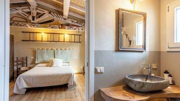 A bedroom with a bed, wooden floor, and a ceiling with exposed beams. There is a bathroom with a stone sink, mirror, and wooden countertop.
