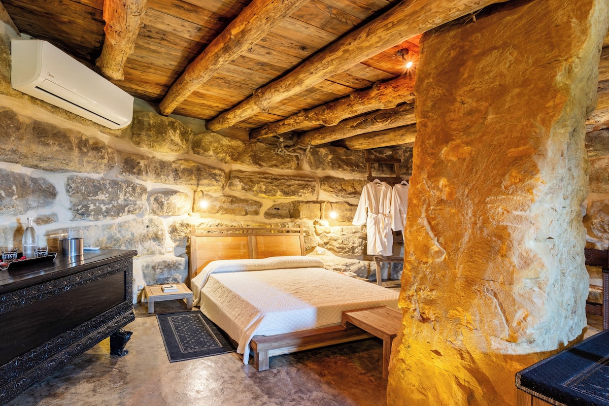 Ein rustikales Zimmer mit Steinelementen, Holzbalkendecke, einem Bett, einer Sitzbank, einem Schmuckkästchen und einem an der Wand hängenden Robe.
