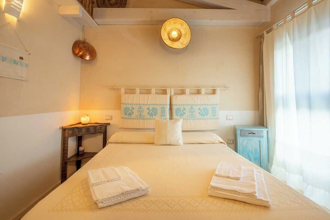 Cama arrumada com toalhas dobradas, mesa de cabeceira com abajur e um pequeno armário.
