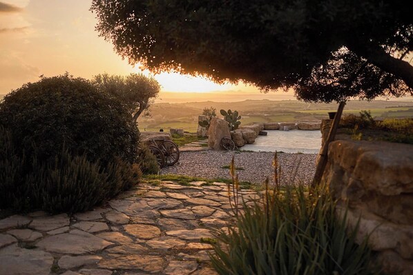 Un sendero de piedra conduce a un puente rústico sobre un arroyo, rodeado de follaje y una vista al atardecer.