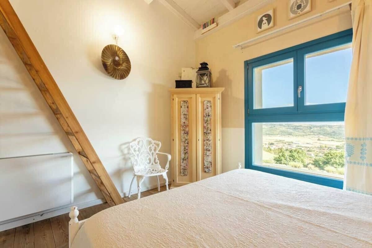 Ein Schlafzimmer mit großem Fenster, einem Bett, einem weißen Stuhl und einem Holzkasten.