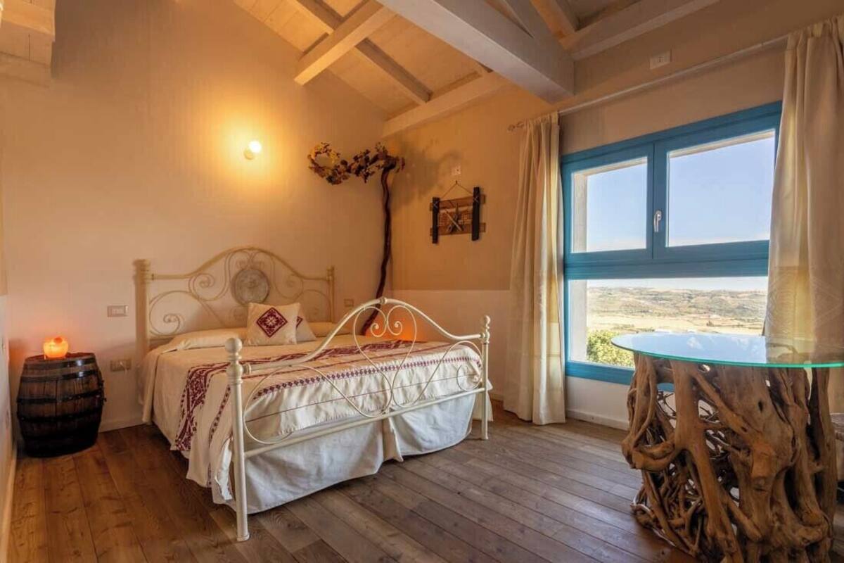 Ein Schlafzimmer mit einem Bett, einem Holzfass, einem Tisch mit Glasplatte und Blick auf die Landschaft durch ein Fenster.