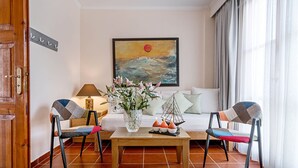 Un salon avec une table basse en bois, deux fauteuils, un canapé, un tableau, un vase de fleurs et une lampe.