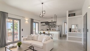 A modern living room with a white sofa, a coffee table, and a kitchen in the background. - BLUEBELL VILLA SUNRISE (ΚΑΜΠΟΣ)