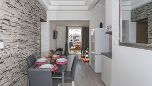 A modern kitchen with a stone wall backsplash, a dining area with a glass table and chairs, and a view of the outside through a doorway. - Apartments Bruno (Okrug)