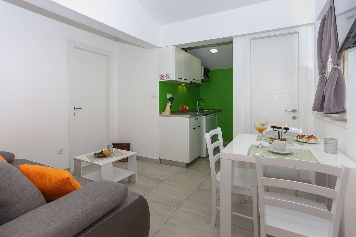 A compact living space with a kitchenette, a dining area with a table and chairs, and a sofa with orange cushions.