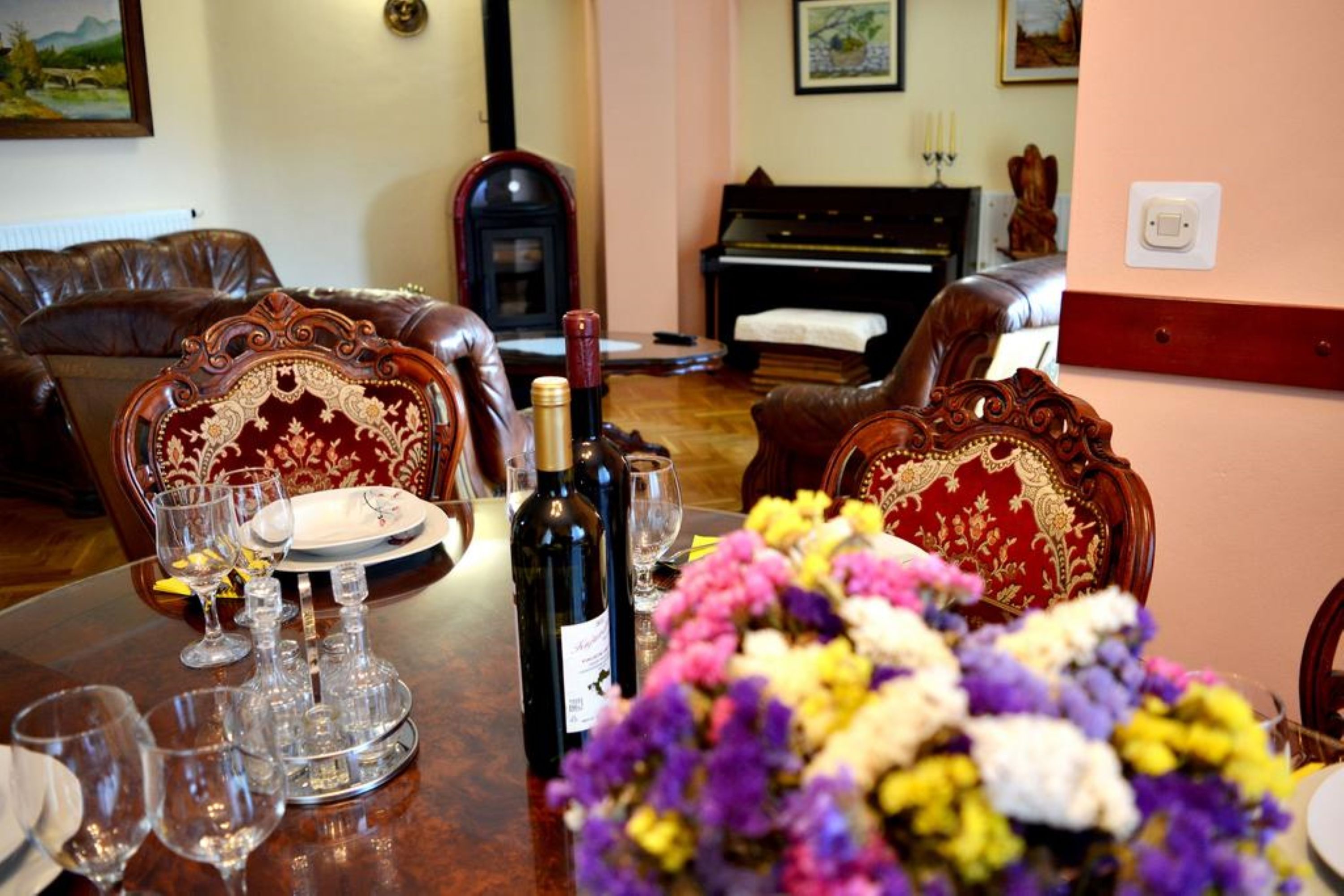 Uma área de refeições com uma mesa de madeira posta para uma refeição, cercada por cadeiras elaboradas. Há uma garrafa de vinho e copos na mesa, e um buquê de flores em primeiro plano.