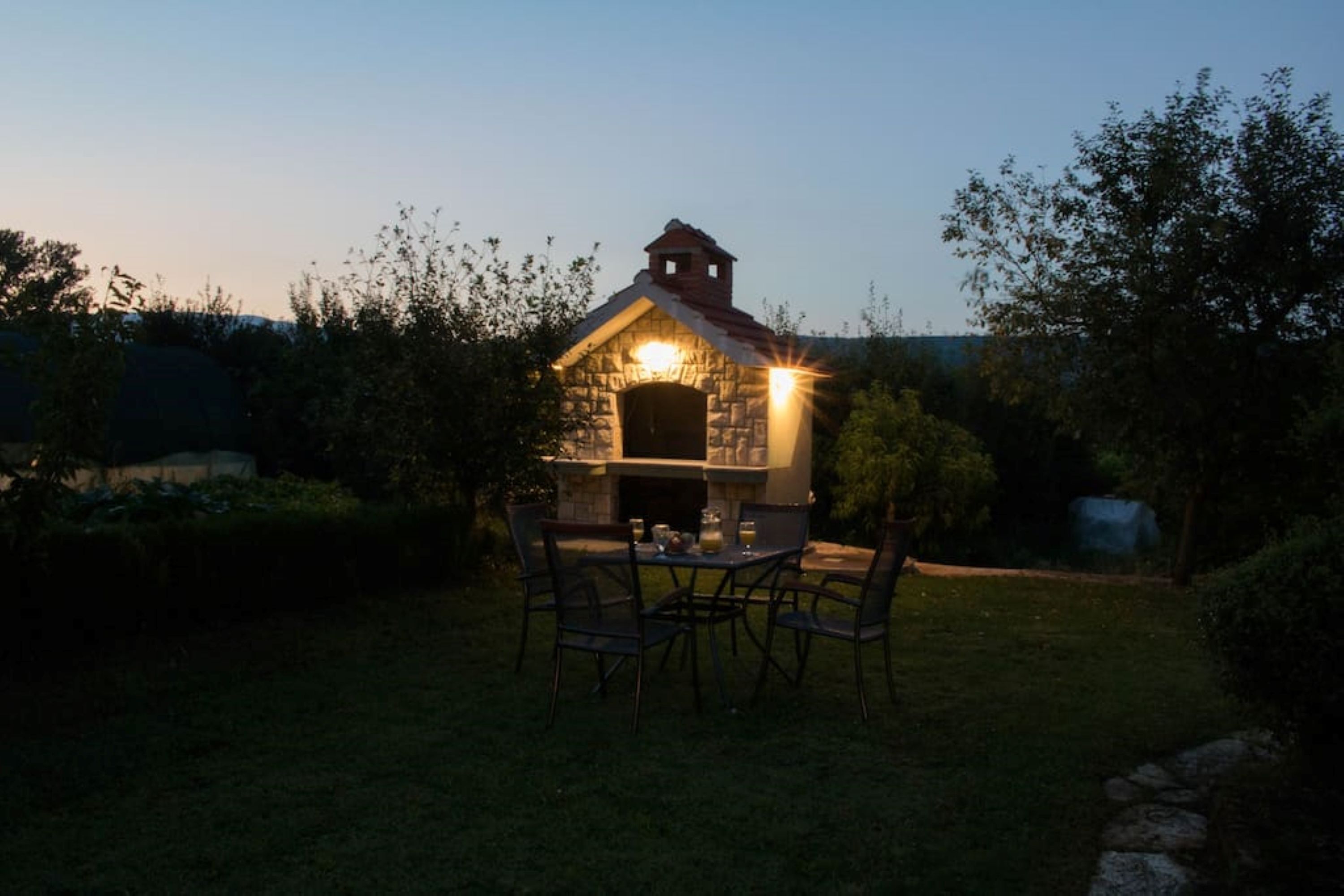 A stone-built outdoor kitchen with a chimney, surrounded by a grassy area and trees.