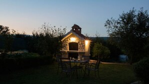 Une cuisine extérieure en pierre, dotée d’une cheminée, entourée d’une pelouse et d’arbres.