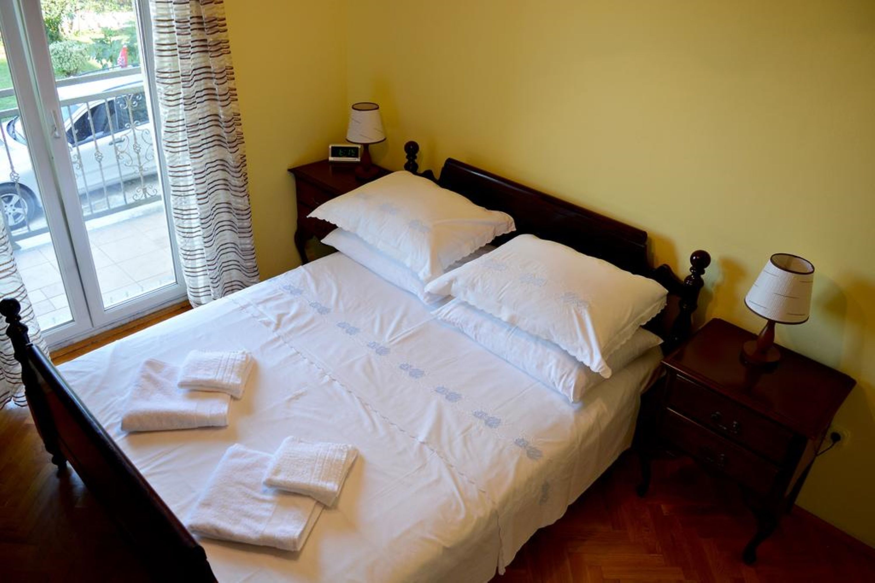 A neatly made bed with white linens and pillows, a bedside table with a lamp, and a view of the outside through a window.